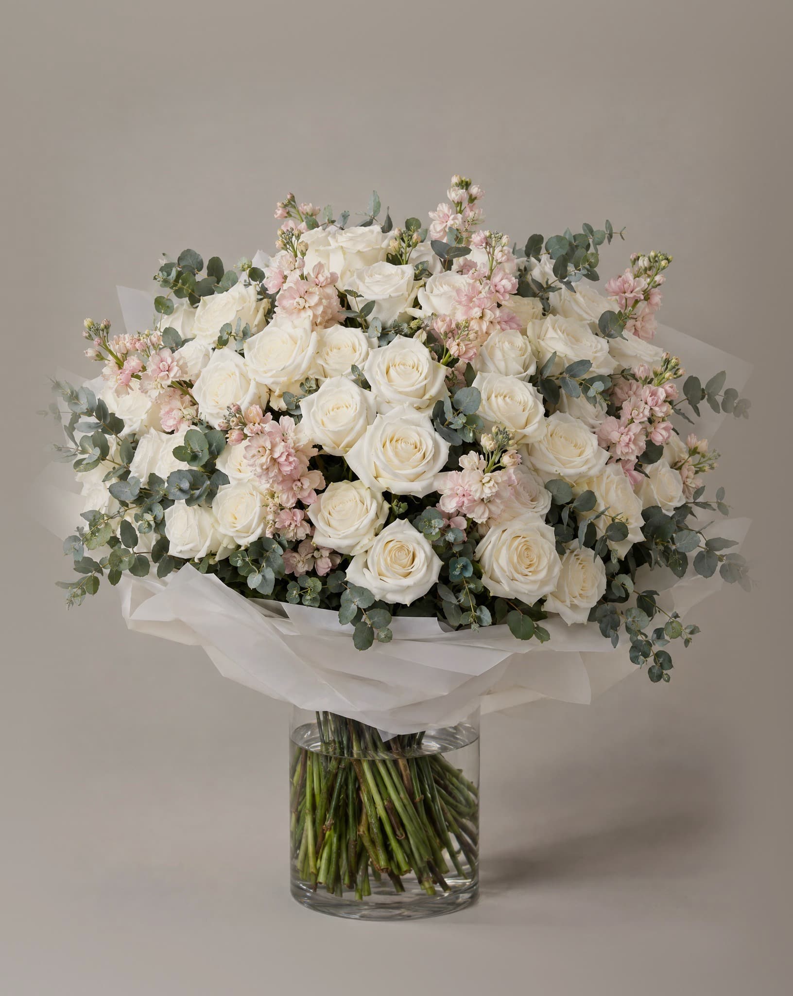 One hundred white roses with pale pink stock and eucalyptus in a wide domed bouquet in a clear glass vase