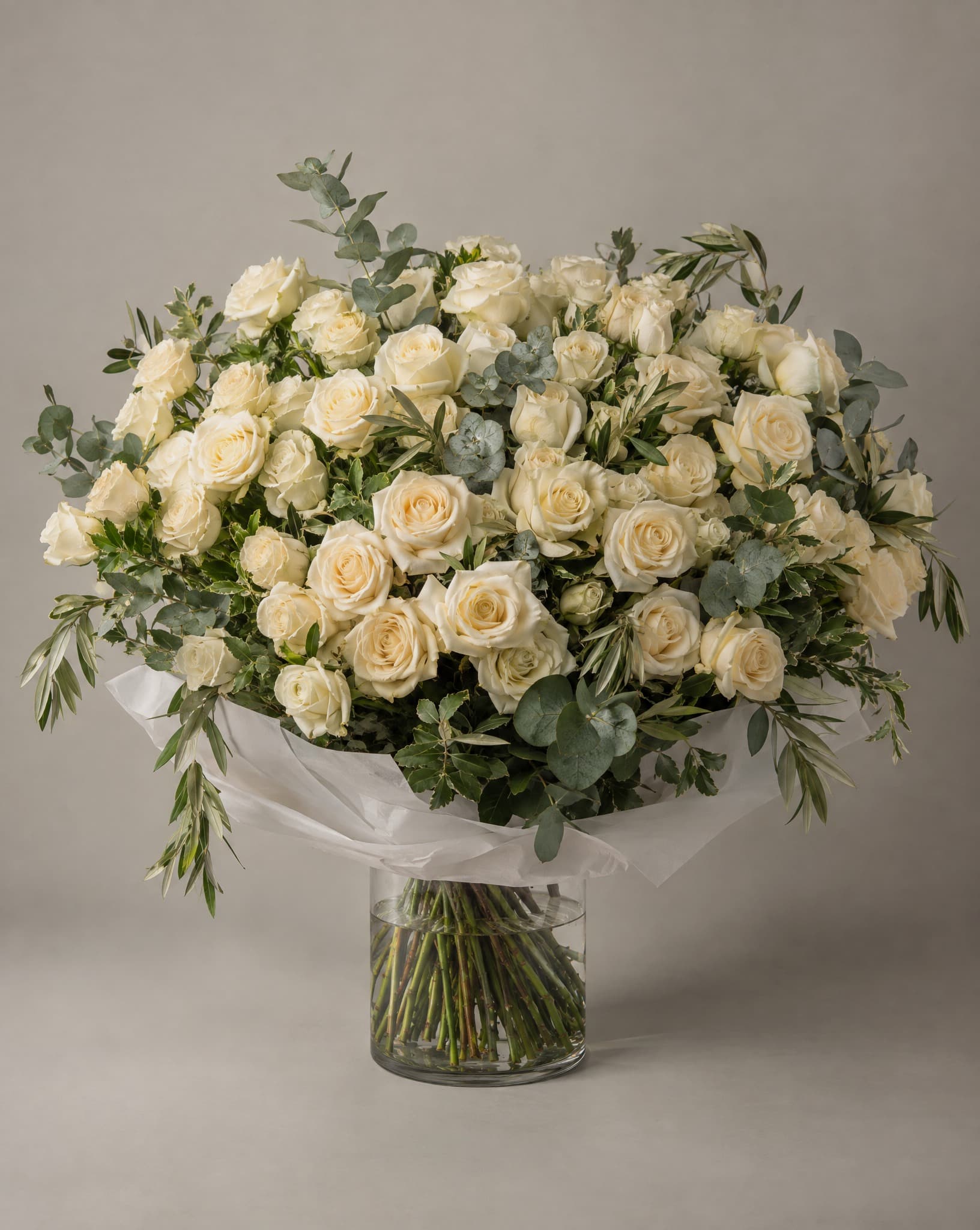 One hundred cream and ivory roses with eucalyptus and olive in a wide, loose bouquet wrapped in white tissue in a clear glass vase