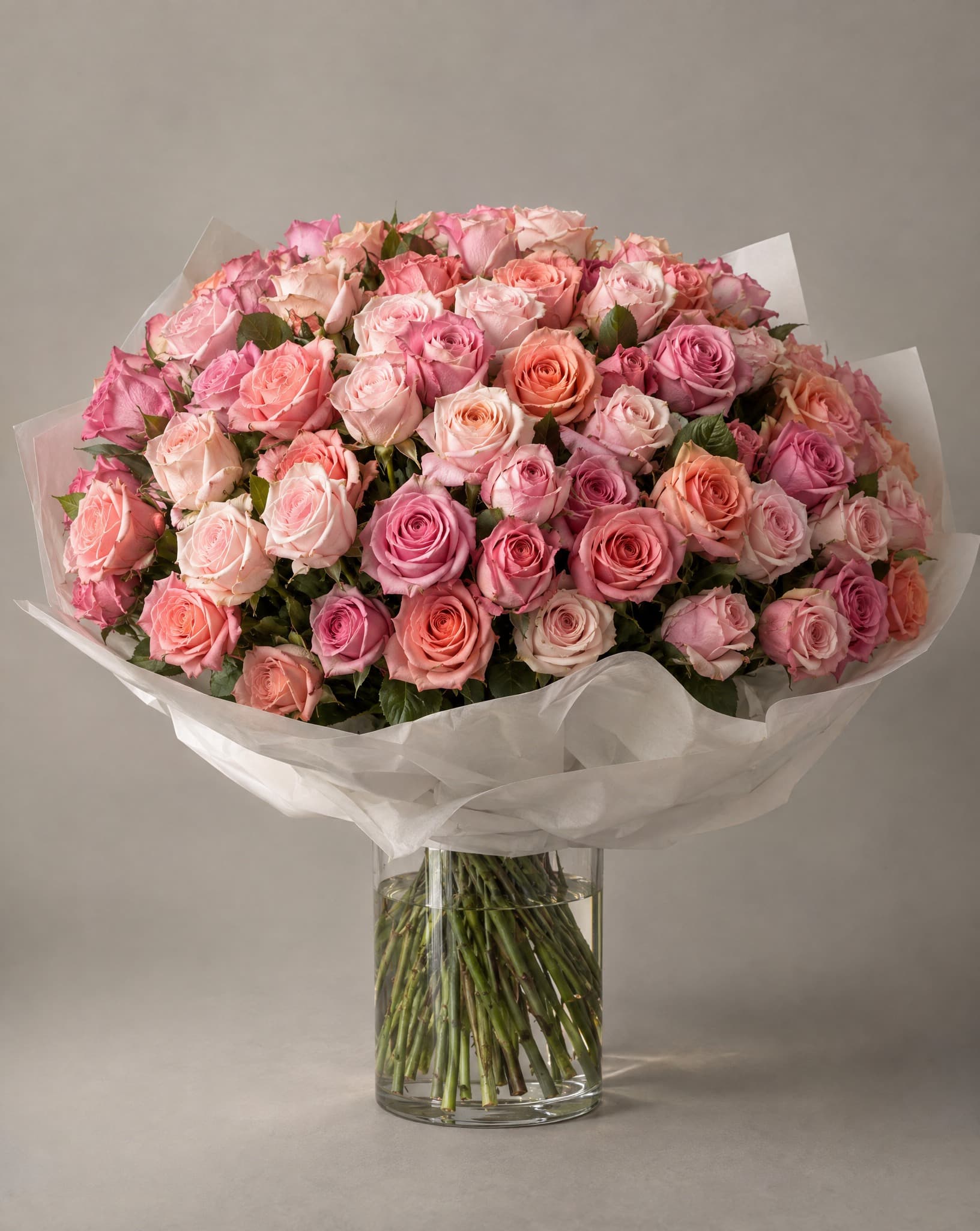 One hundred pink, coral and cream roses in a domed bouquet wrapped in white paper in a clear glass vase